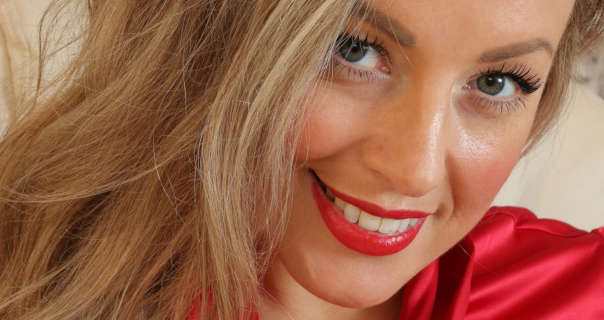 Brianna Olivia is seen in a bright red lipstick with her light blonde hair loose and pushed to one side. She appears in front of a cream wall. Brianna wears a soft red blouse and lash defining mascara.