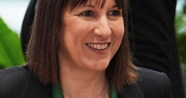 Rachel Reeves smiles widely. She wears a black blazer over a green top. She sits in a blue and green room during a Canary Wharf meeting.