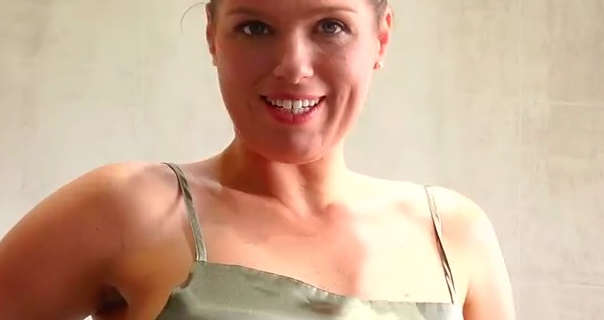 Lottie Lalay is seen smiling and looking into the camera during a SatinSilkFun shoot. she wears a dark sage green dress in a cream tiled bathroom. Lottie has her blonde hair tied up in a bun.