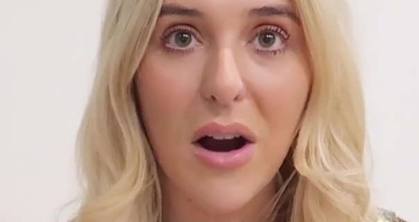 Em Sheldon is seen with her mouth open. She appears in front of a dark white wall. Em wears her blonde hair loose and lightly curled. Her makeup includes a gloss pale pink lipstick and black clumping mascara.