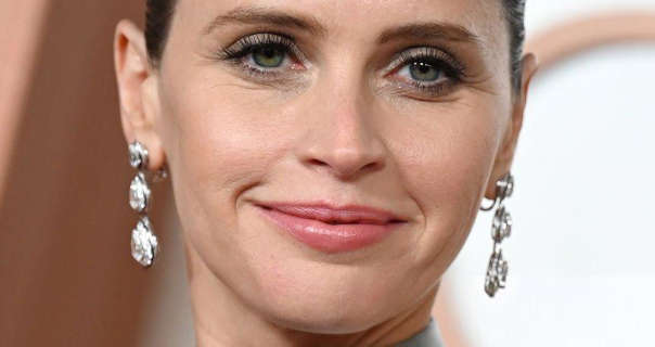 Felicity Jones is seen smiling a little as her picture is taken. She wears a pair of large dangling silver gemstone earrings. Felicity appears with her hair tied up and with a dark pink lipstick and black mascara. She stands in front of a white and rose gold wall.