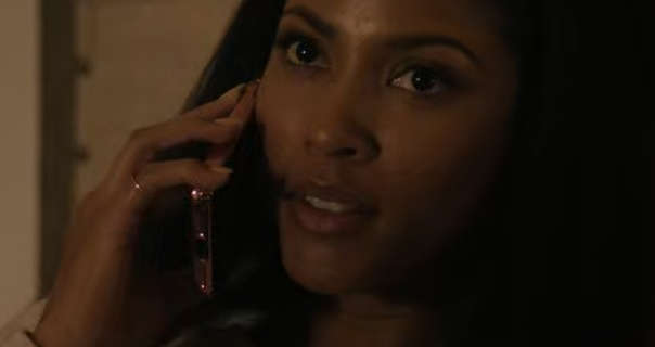 Lex Scott Davis is seen talking on a gold frame mobile phone in a dark room. She wears a matte lipstick as she lies on a large bed. Lex has her thick black hair down and straight.