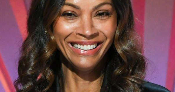 Zoe Saldana laughs and smiles widely. She stands in front of a dark pink and pale purple backdrop. Zoe wears her black hair down, curled and with brown highlights. She is seen in a shiny black top and a dark pink glossy lipstick.